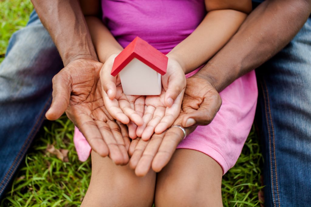 Symbolisch houten huisje in handen van een kind, dat voor een volwassene zit, die hier ook zij handen omheen heeft gevouwen.